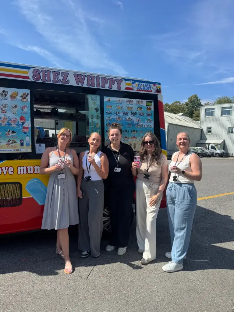 Shez Whippy, Ice Cream van for events. A group of adults pose in front of a Shez Whippy ice cream truck at a corporate event, with the truck’s ice cream and ice-cream menu visible behind them.